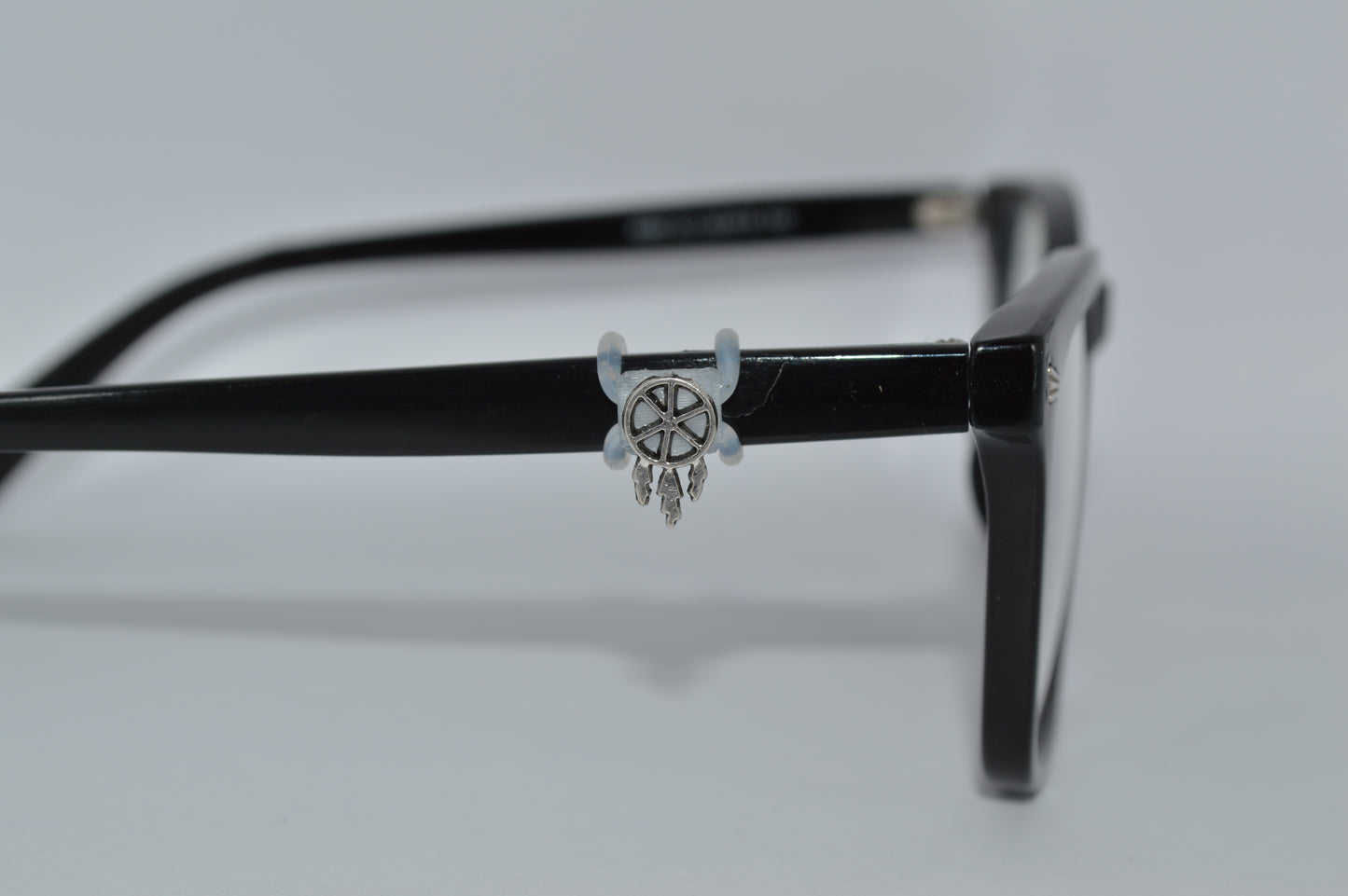 A silver dreamcatcher charm attached to the arm of a pair of black eyeglasses